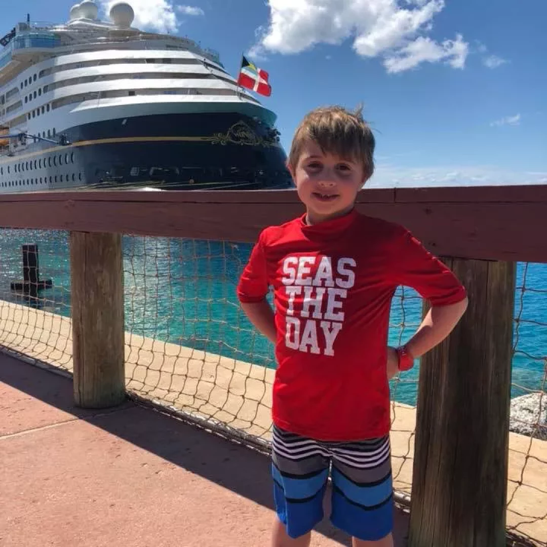 Young boy standing in front of a cruise ship, wearing a red shirt that says SEAS THE DAY