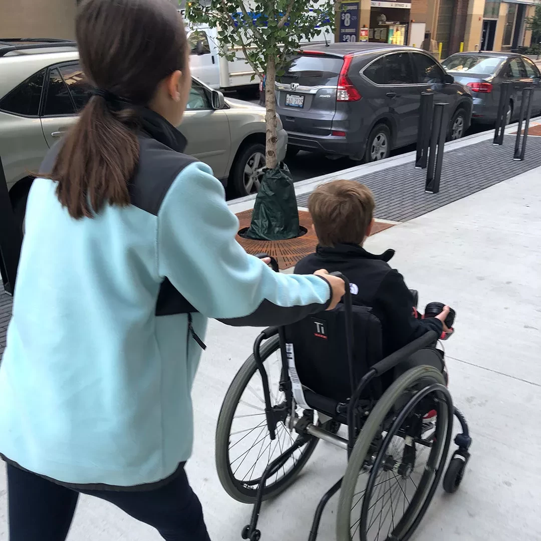 Person pushing a young boy in a wheelchair along a city sidewalk