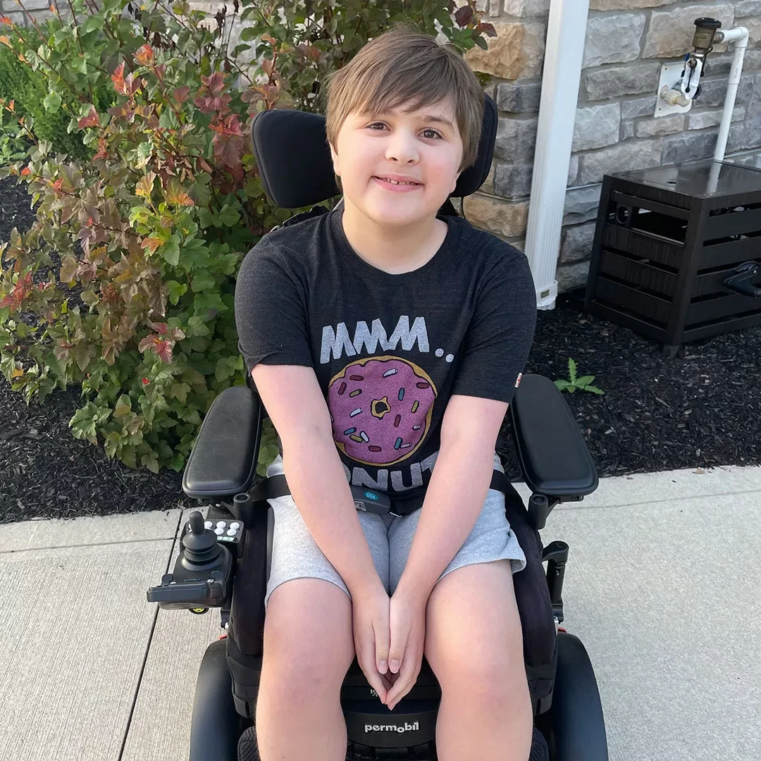 Smiling young boy sitting in a motorized wheelchair, wearing a t-shirt with a donut graphic.