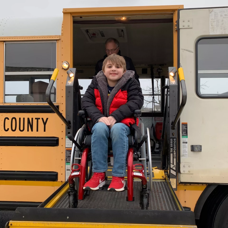 Young boy in a wheelchair smiling as he is lifted onto a school bus.