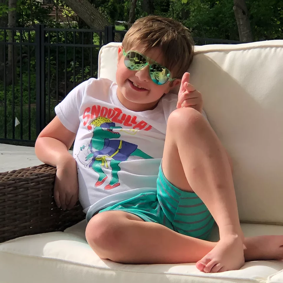 Young boy relaxing on an outdoor couch, wearing sunglasses and a 'Snoozilla' t-shirt, giving a thumbs-up.