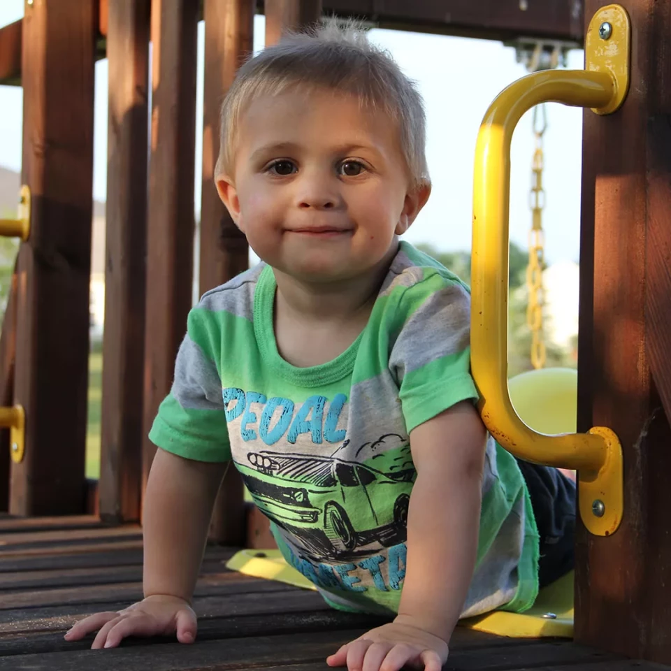 Young boy smiling while playing on a playground, wearing a green shirt with a car graphic.