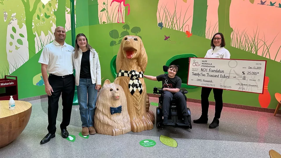 Group of people presenting a $25,000 donation check to a foundation, standing next to large dog statues, with Alex in a wheelchair smiling.